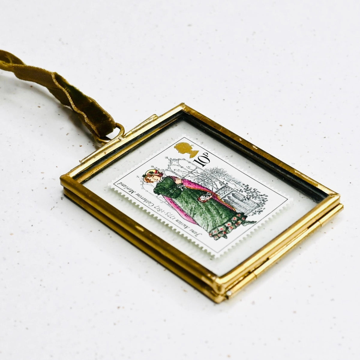 A limited edition commemorative stamp featuring Catherine Morland, mounted in a brass hanging frame with a velvet ribbon, laying flat on a speckled, white bakground.