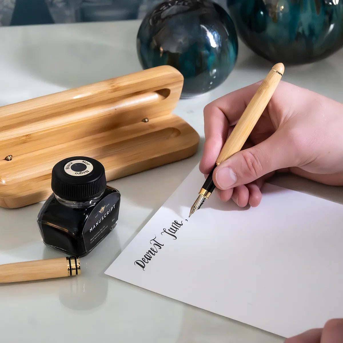 Person writing with a wooden pen on paper, next to an inkwell and fountain pen parts.