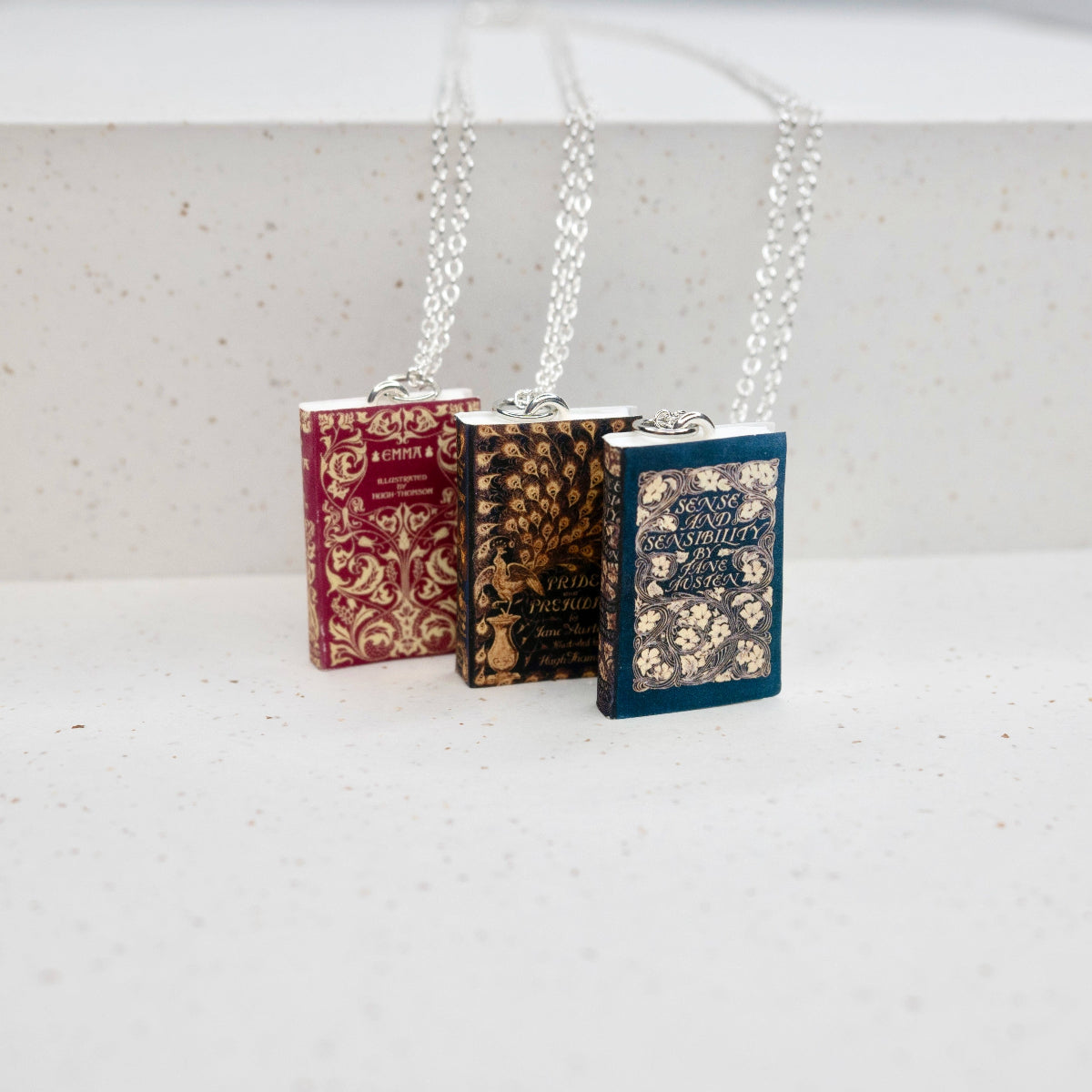 Three book necklaces are in a row against a speckled, white backdrop. The books are Emma, Pride and Prejudice, and Sense and Sensibility.