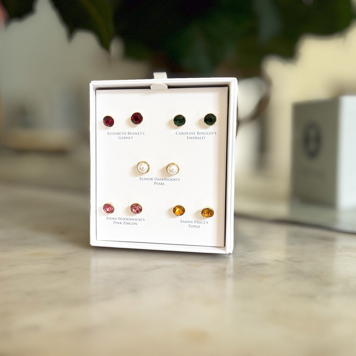 White Jane Austen branded box with five pairs of stud gemstone earrings with character names attributed.