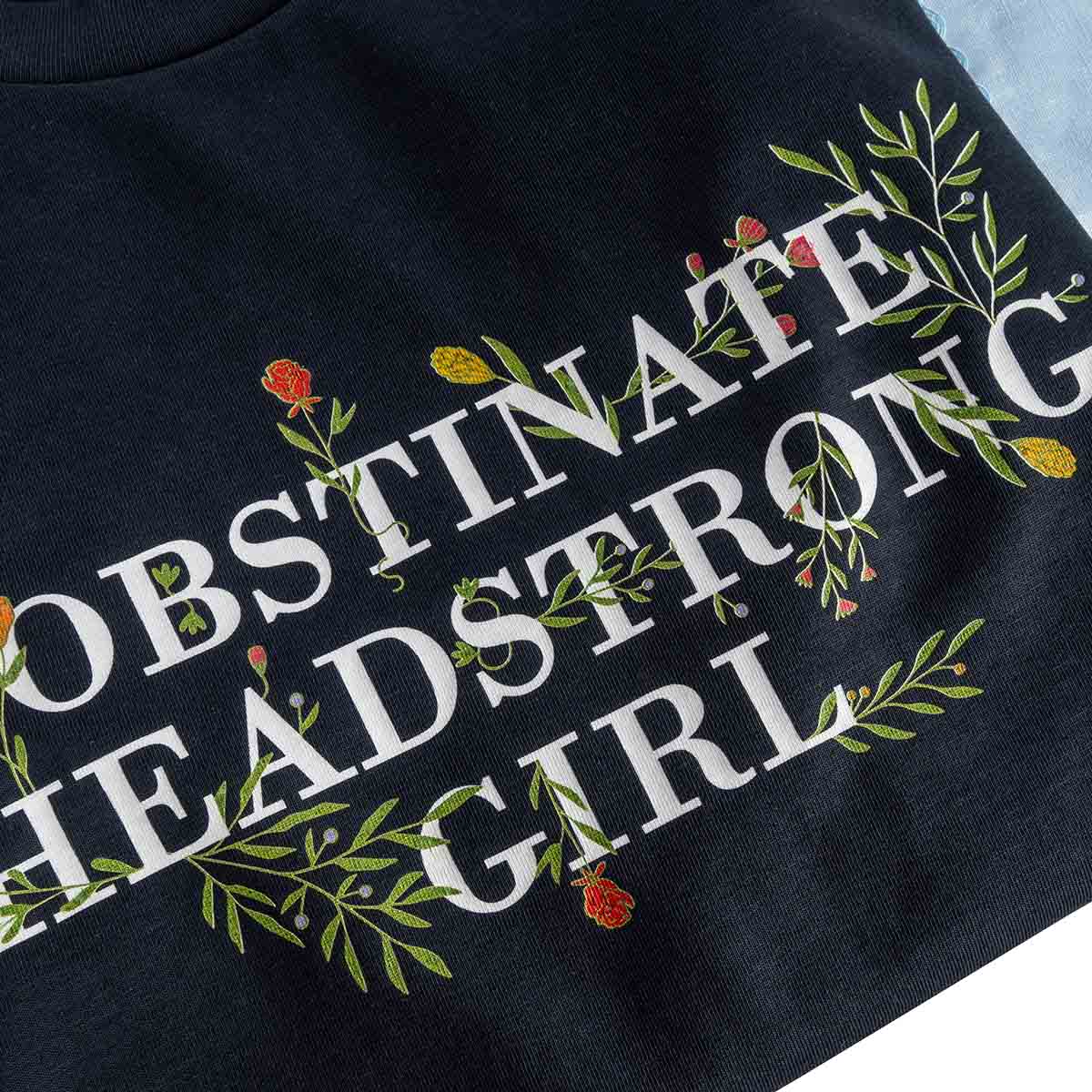 Black shirt with white text 'OBSTINATE HEADSTRONG GIRL' and floral designs on a light blue background