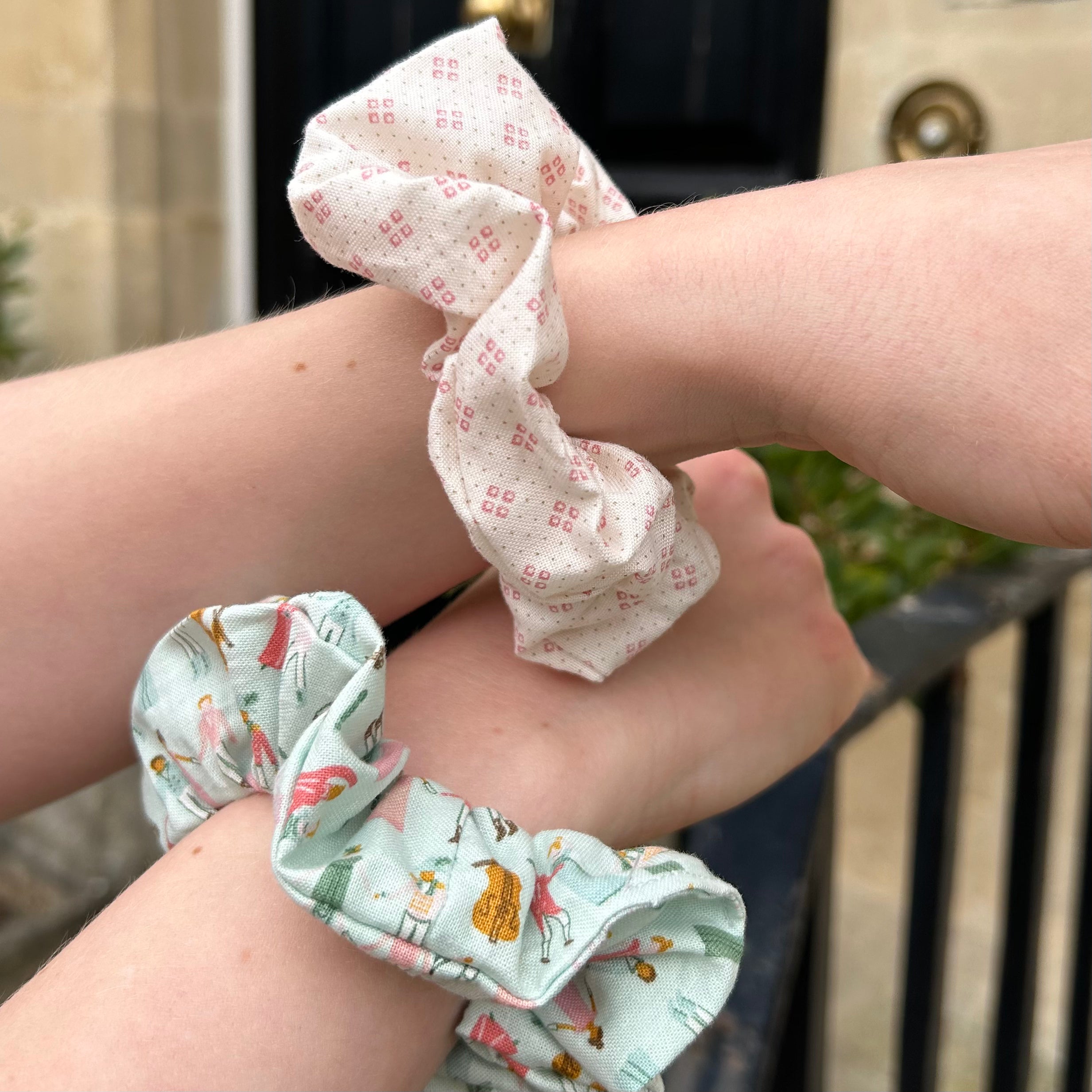 Regency Ball Scrunchie (pale blue main colour with people dancing) with a pink patterned scrunchie shown on wrists.