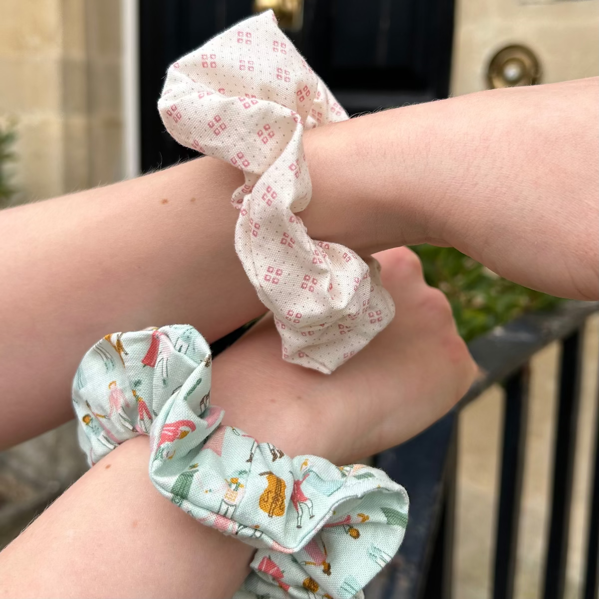 Regency Ball Scrunchie (pale blue main colour with people dancing) with a pink patterned scrunchie shown on wrists.