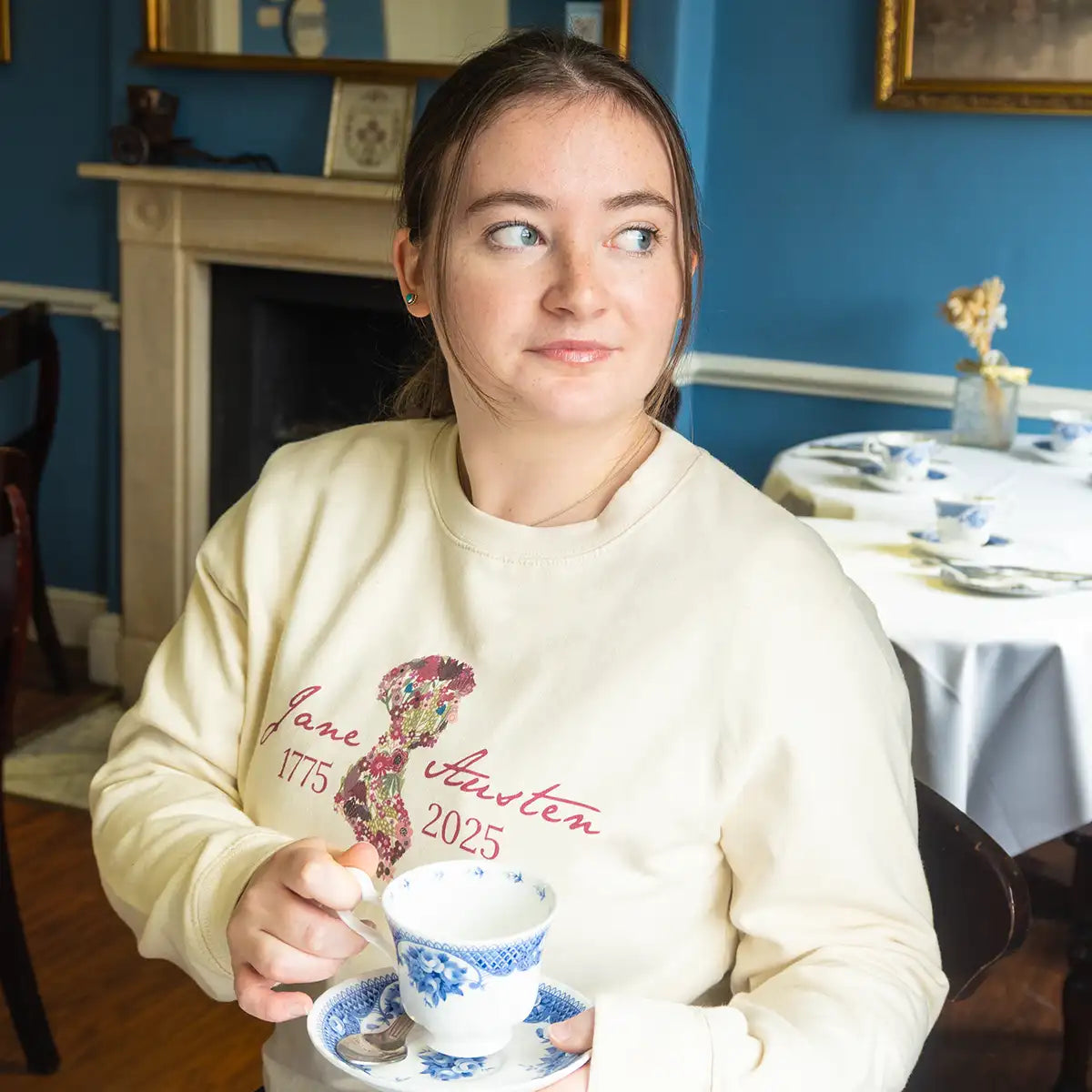 Model wearing a cream Jane Austen-themed sweatshirt holding a teacup in the Jane Austen Centre's Regency Tea Room.