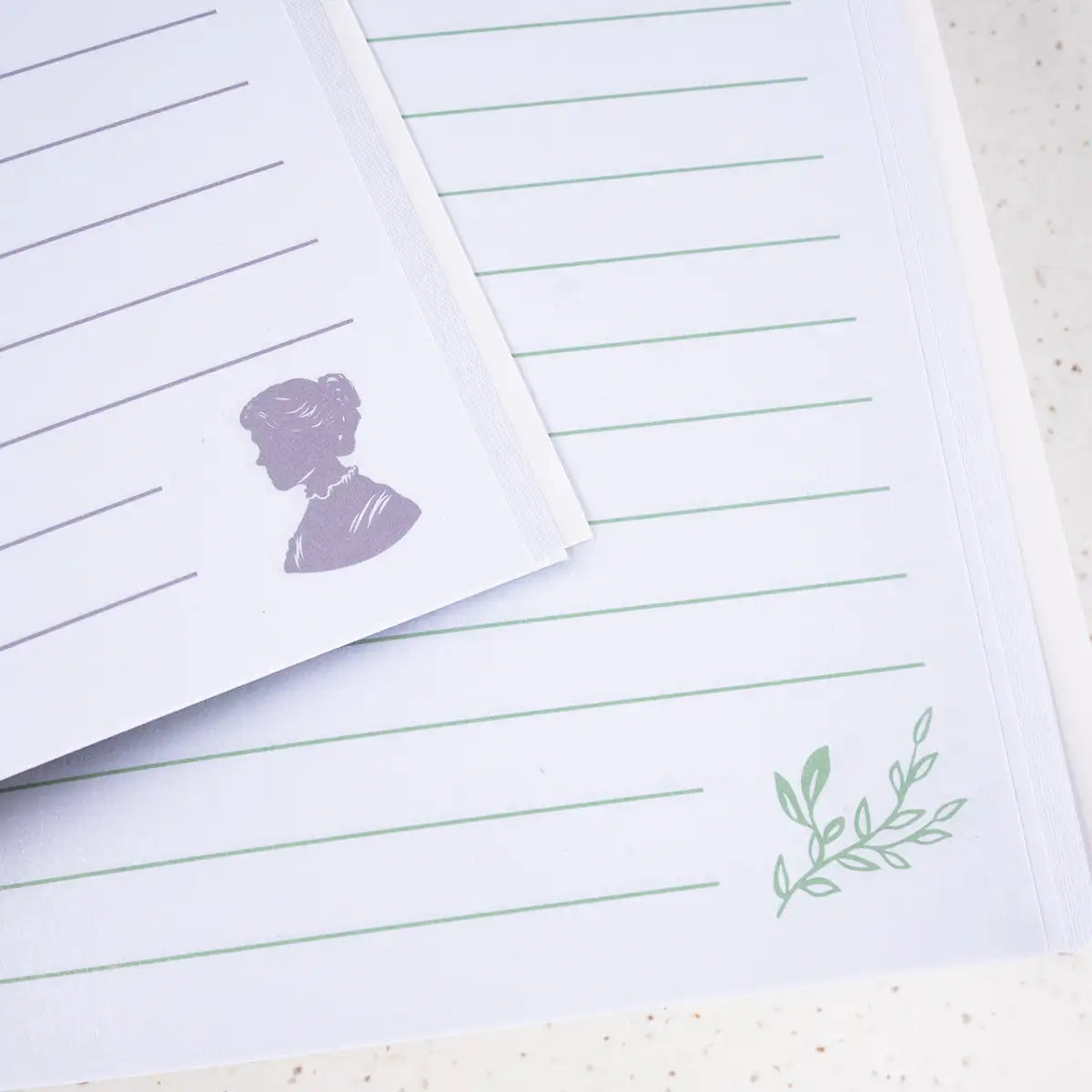 Two sheets of lined paper with a purple silhouette on one and green leaf design on the other. Photographed on a textured surface.