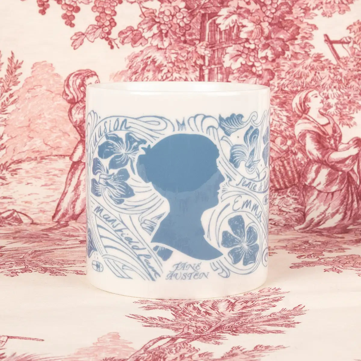 Front view featuring Jane Austen silhouette in blue. Cream mug decorated in swirling blue flower illustrations and the names of Jane Austen's novels. Gold handle.