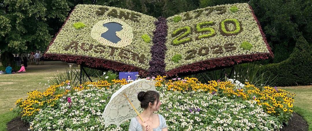 Woman in a white dress sitting in front of a floral display shaped like an open book with '250' on it, surrounded by trees and flowers.