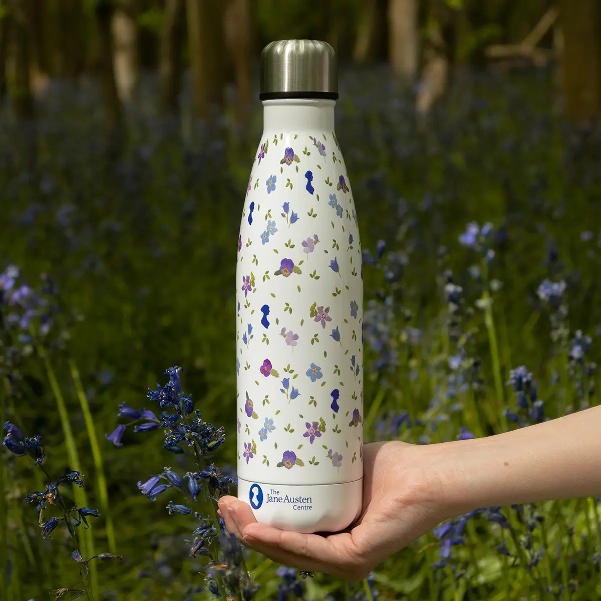 A hand holding a floral water bottle in the woods above some bluebells