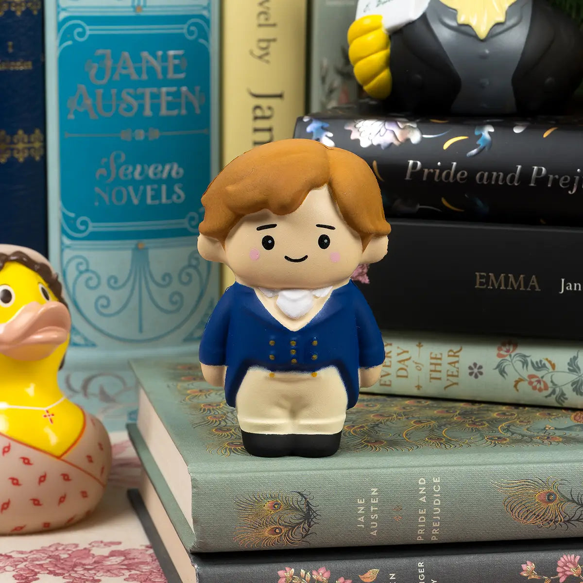 Mr Darcy Squishi Pal foam stress ball in blue regency attire, on a stack of books written by Jane Austen, surrounded by regency themed rubber ducks with more books written by Jane Austen stacked up and lined up behind him