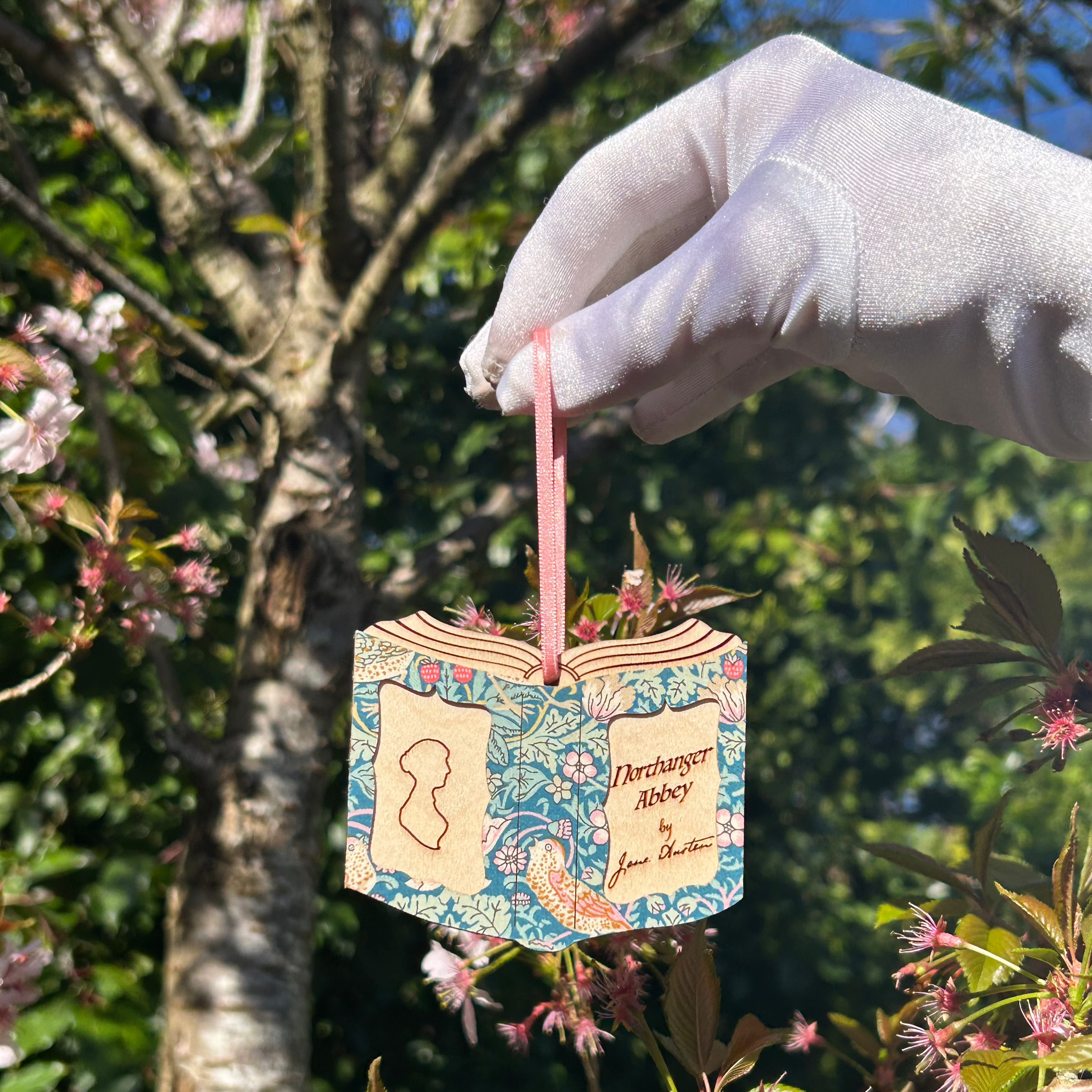 Surrounded by cherry blossoms and leaves in golden sunlight, a satin gloved hand holds this wooden Northanger Abbey decoration by a pink ribbon.