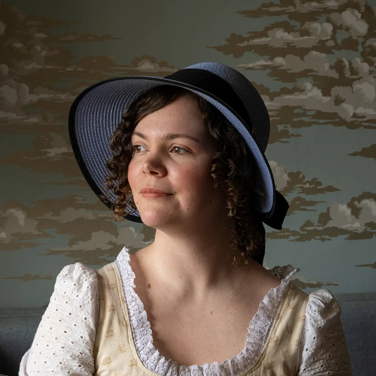 A model in a regency dress wearing a blue open-back regency bonnet against a blue background with gold clouds