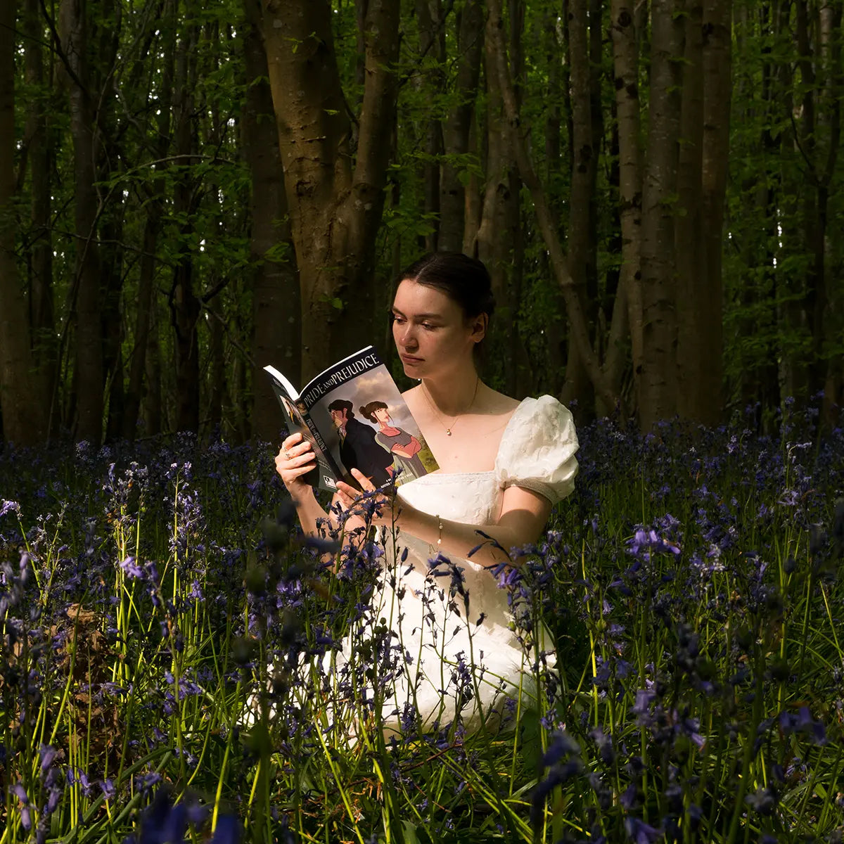 A woman sitting in a patch of bluebells in the woods, reading the Pride and Prejudice graphic novel
