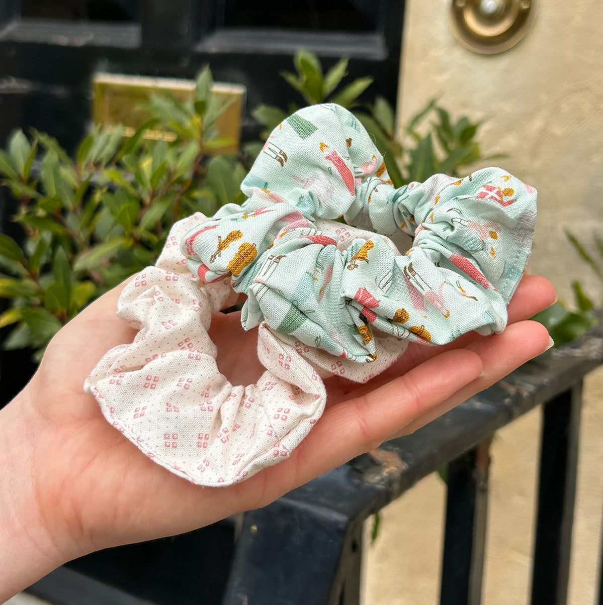 Regency Ball Scrunchie (pale blue main colour with people dancing) with a pink patterned scrunchie in the palm of hands.