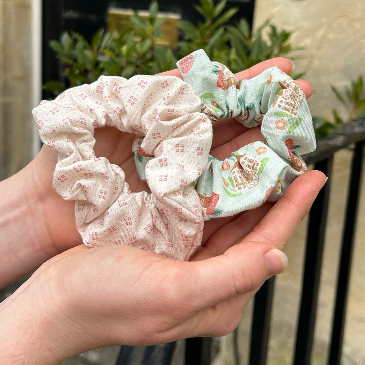 Regency House Scrunchie (Pale blue main colour and floral pattern) with a pink patterned scrunchie in the palm of hands.