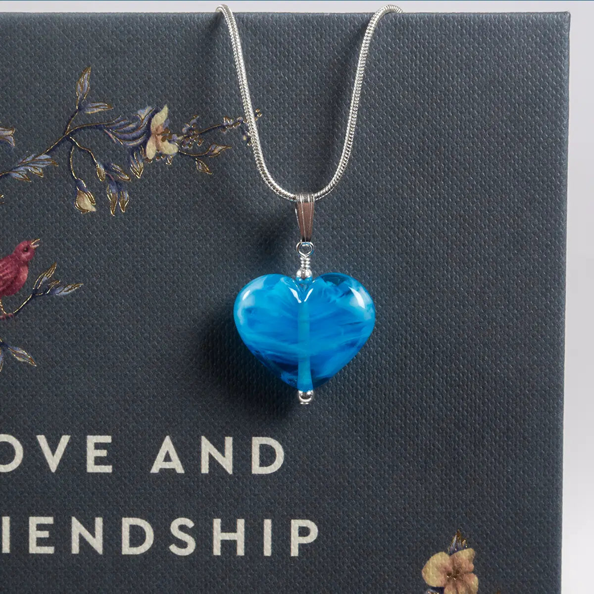 a blue glass heart on a silver chain hanging on a book cover of jane austen's love and friendship