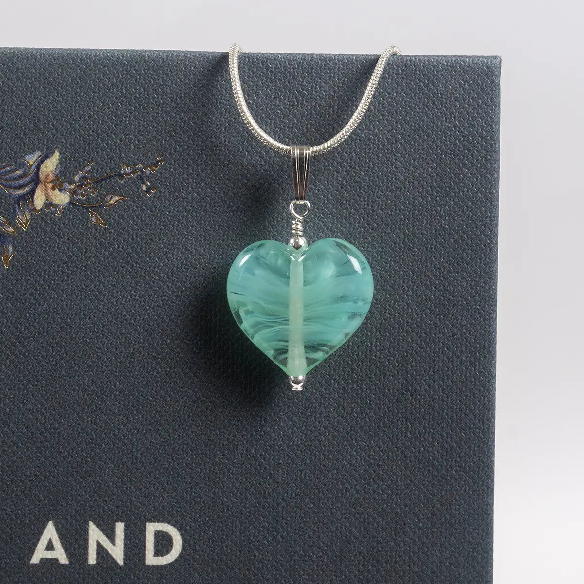 a mint coloured glass heart necklace pendant on a thin silver chain hanging from a dark grey book cover against a white background