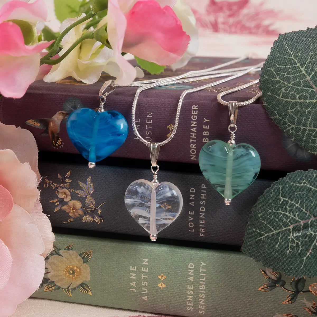 Three different coloured glass necklaces with silver chains, on a stack of books, surrounded by artificial flowers