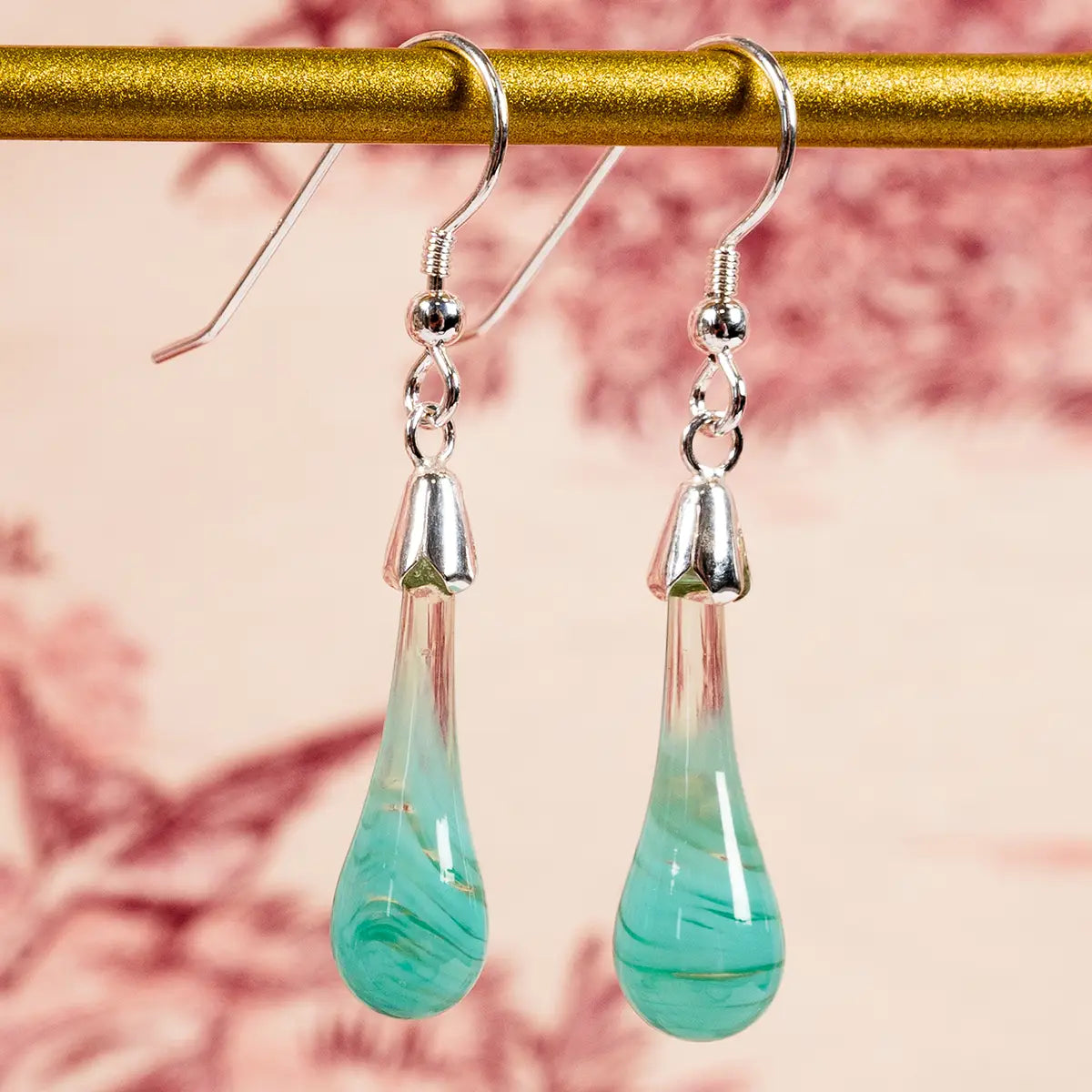 mint coloured glass drop earrings with silver detailing hanging from a gold bar against a cream and red patterned background