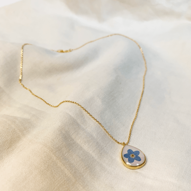 The necklace is laid out on a soft cotton sheet to show it's full beauty. The 14ct gold plated chain has the pendent hanging on one side, and a lobster claw clasp on the other. The pendent is a tear drop shape with a milky white colour and a forget me not in the middle. The pendent is rounded with 14ct gold plating to define it's delicate shape. Jane Austen Jewellery Regency Jewellery
