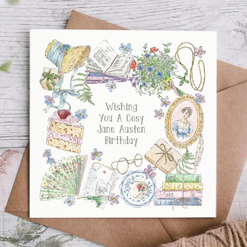 "wishing you a cosy jane austen birthday" card shown with the brown envelope behind. The card has a collection of green foliage, pretty pink cake, colourful fan and a traditional image of Jane Austen.