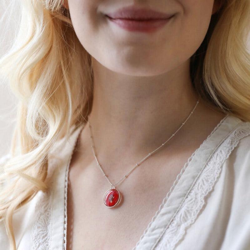 A woman wears the December necklace. The necklace features a strong red flower set in resin, hanging from a silver satellite chain.