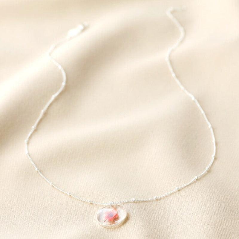 The June necklace is laid out on a silk backgrop. The satellie chain is splayed out to show its 16 inches and the dainty red-pink flower is set in resin as a pendant