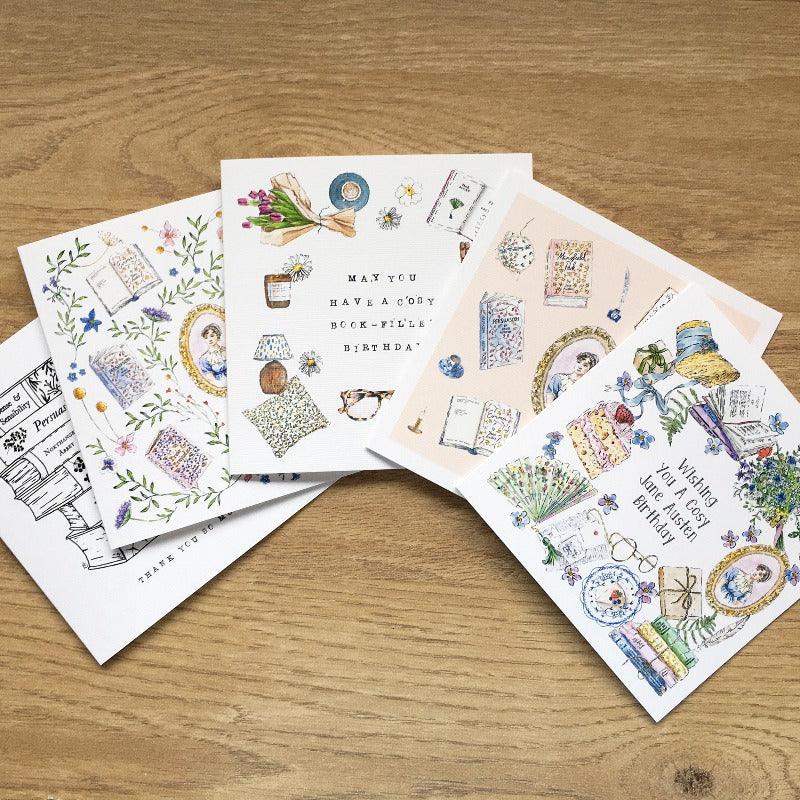 the cards lay out of a wooden table, displaying each pride and prejudice and jane austen greetings card.