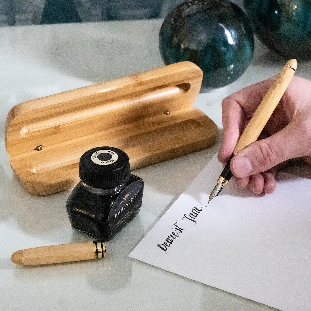 Person writing with a wooden pen on paper, next to an inkwell and fountain pen parts.