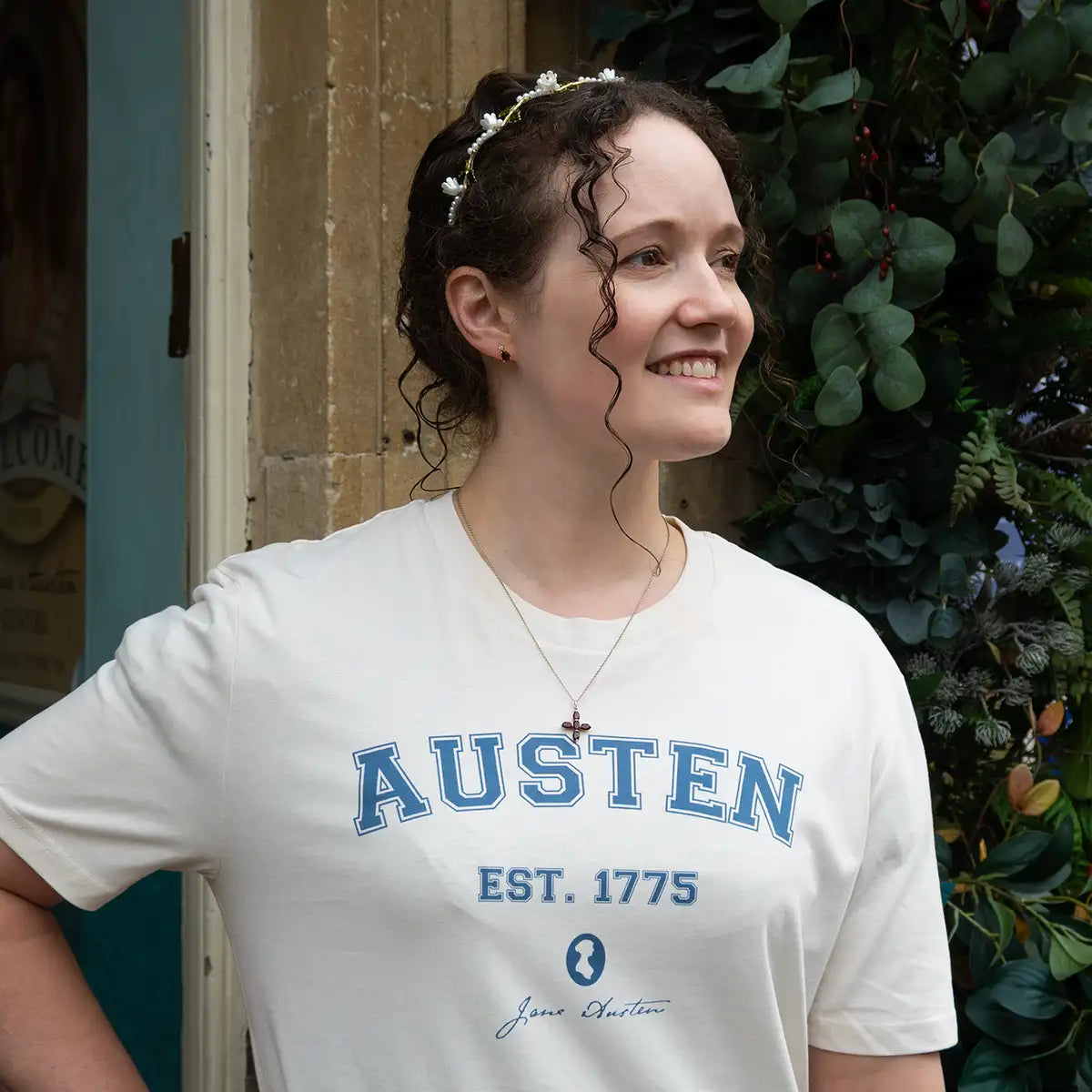 Woman in beaded headband outside wearing a cream shirt with 'AUSTEN EST. 1775' and Jane Austen silhouette printed in blue. 