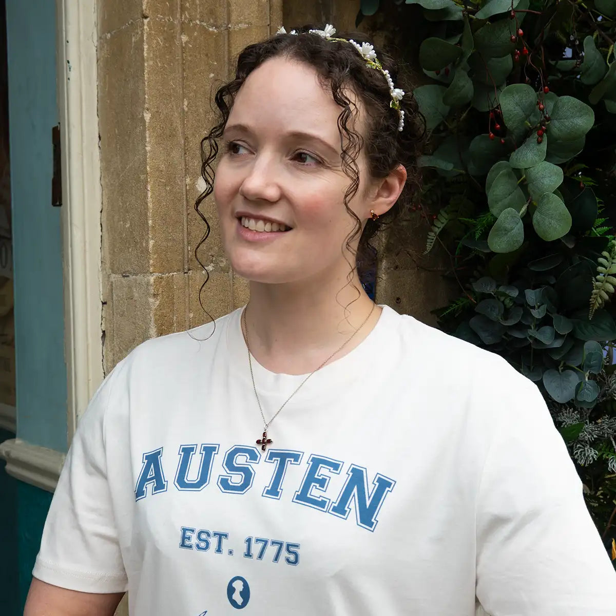 Woman in beaded headband outside the Jane Austen Centre wearing a cream shirt with 'AUSTEN EST. 1775' and Jane Austen silhouette printed in blue. 