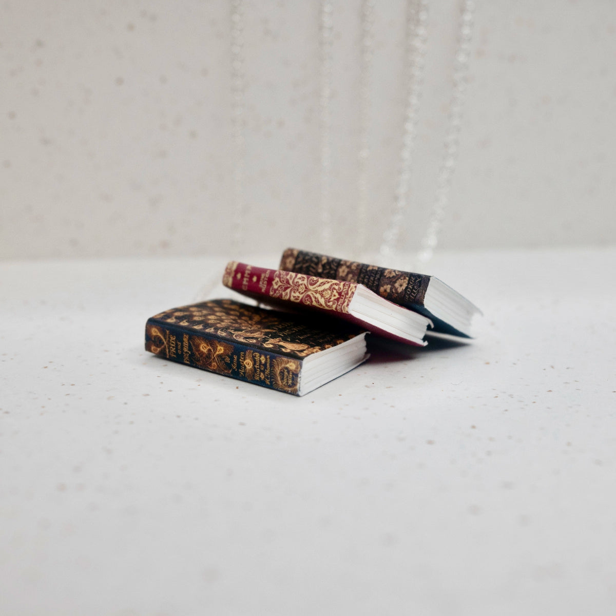 Three book necklaces form a collapsed pile, the "pages" are visible. They are arranged against a speckled, white backdrop.