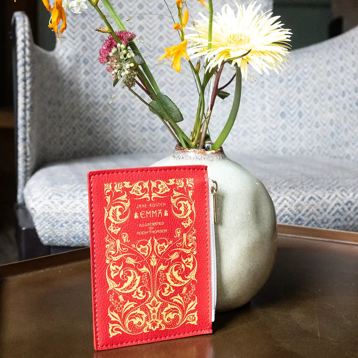 Decorative vase with flowers behind a red coin purse titled 'Emma' by Jane Austen on a wooden surface.