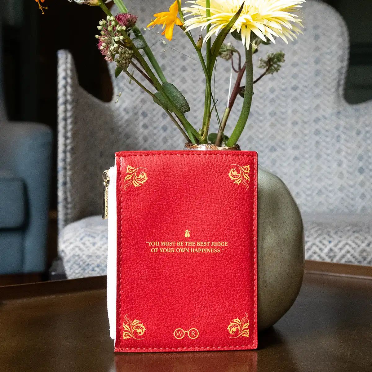 Red coin purse with gold text reading 'You must be the best judge of your own happiness' and floral arrangement behind on a table.