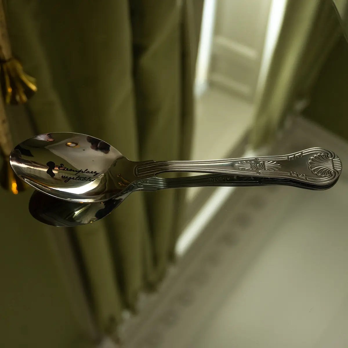 metal spoon with 'exemplary vegetables' printed on it, placed on a mirrored surface.