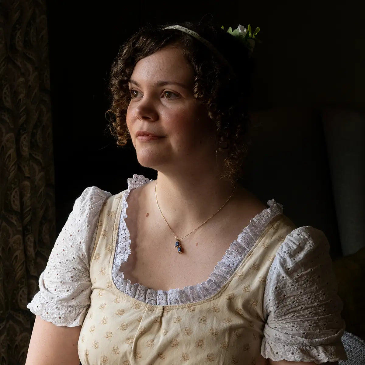 Woman in a historical dress with bluebell necklace and  lace details against a dark background
