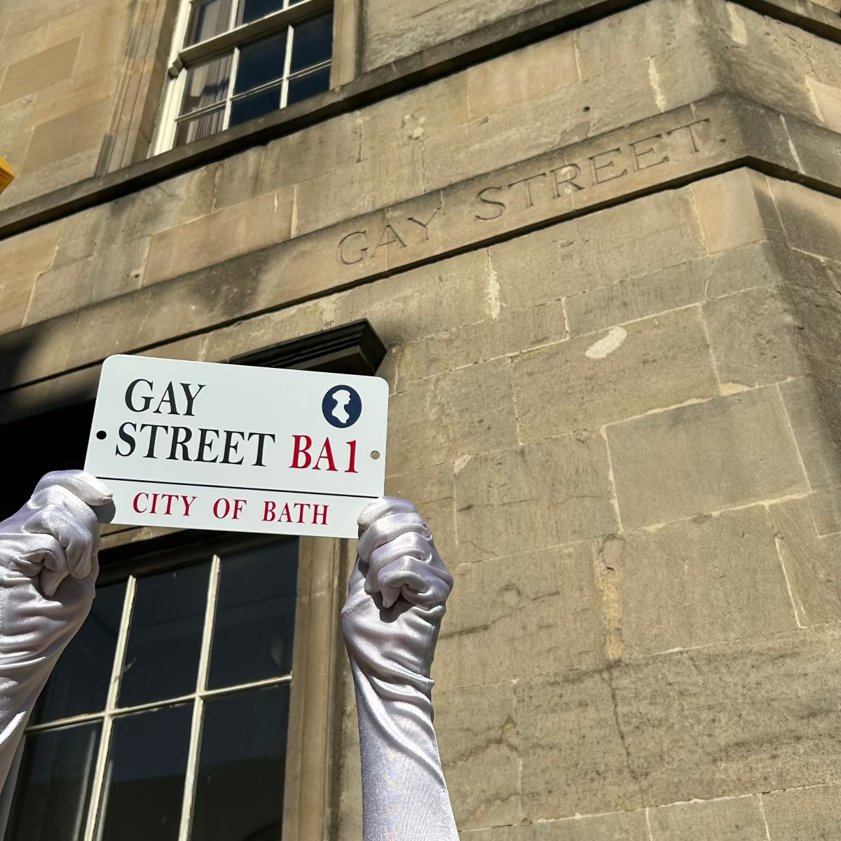 Gloved hands hold up a metal Gay Street Sign in front of a sandstone building, carved with the words Gay Street.