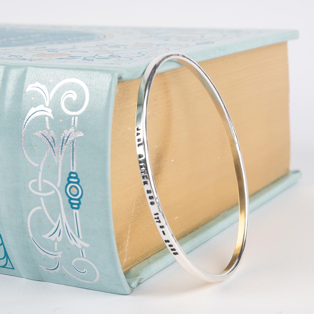 Silver bangle bracelet engraved with 'Jane Austen 250 1775-2025' leaning against a book.