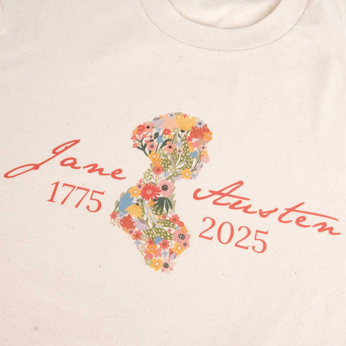 Close-up. Jane Austen 250th Anniversary T-Shirt in Cream. Silhouette of Jane is filled with flowers in warm tones and text 'Jane Austen 1775-2025' is in a warm orange.