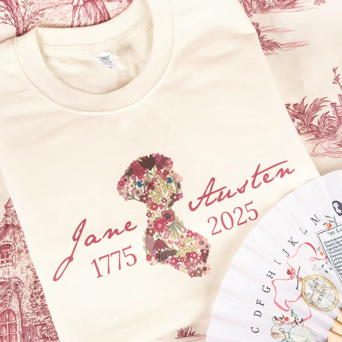 Cream sweatshirt with burgundy floral design and text, surrounded by decorative items on a light background.