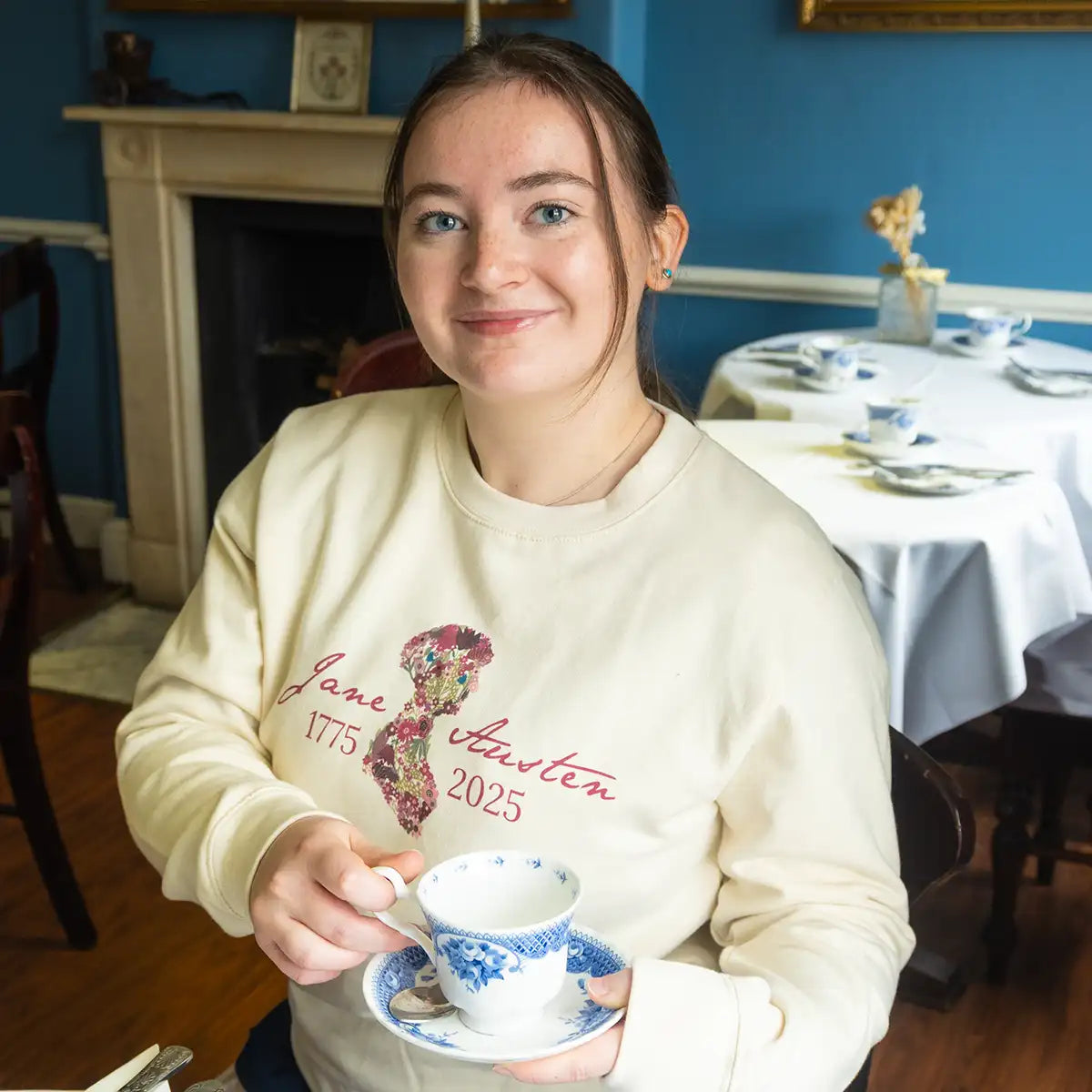 Model wearing a cream Jane Austen-themed sweatshirt holding a teacup in the Jane Austen Centre's Regency Tea Room.