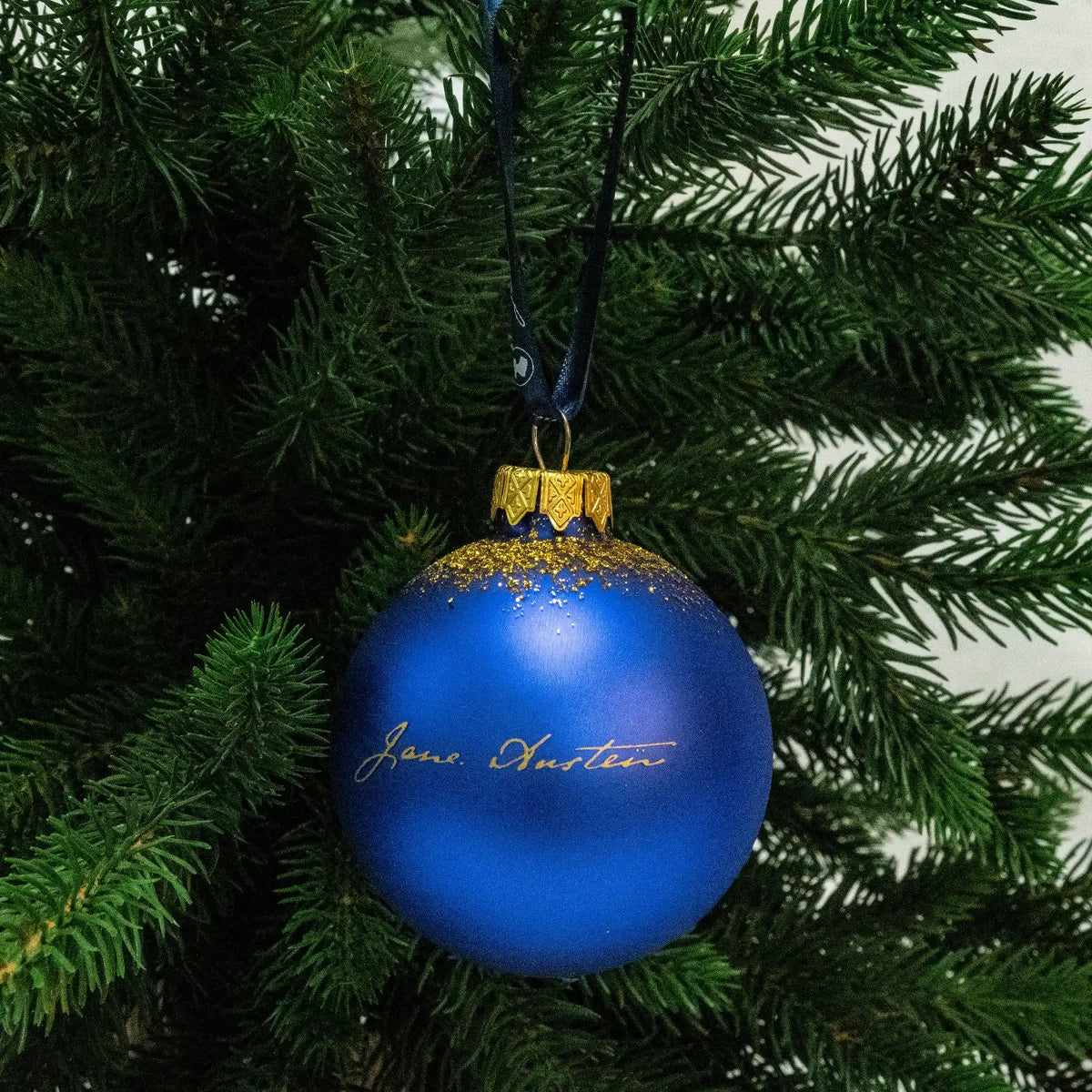 Blue bauble featuring Jane Austen's signature in gold, it is hanging on a Christmas tree from a blue, Jane Austen branded ribbon.