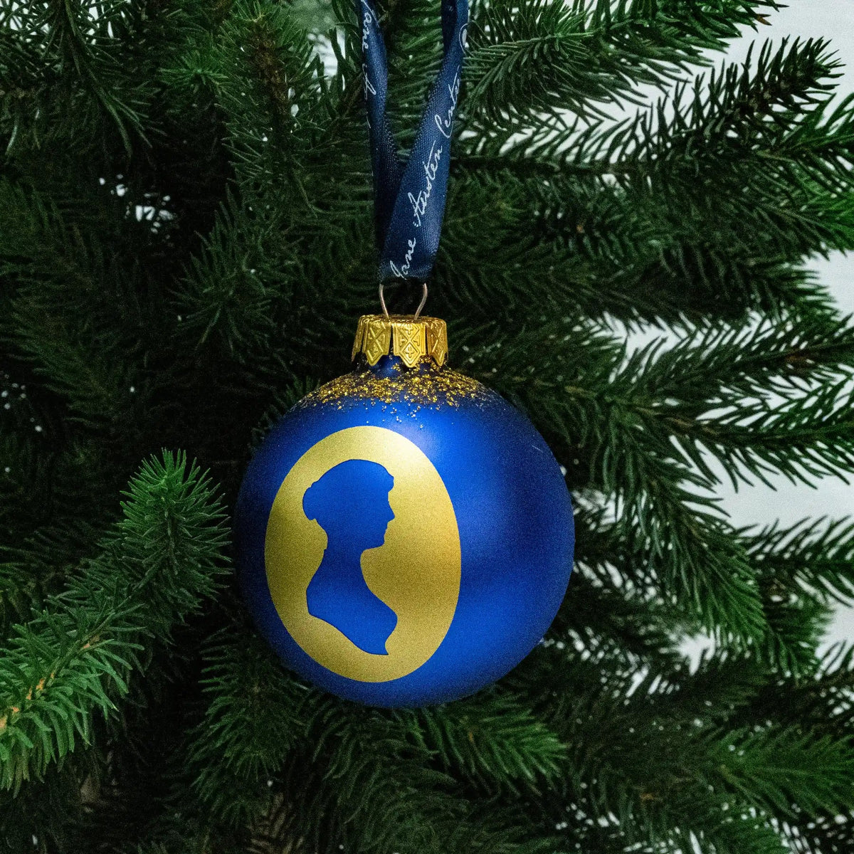 A blue bauble featuring a gold oval from which the silhouette of Jane Austen has been removed, showing the colour of the bauble. It is hanging in a Christmas tree from a blue, Jane Austen branded ribbon. 