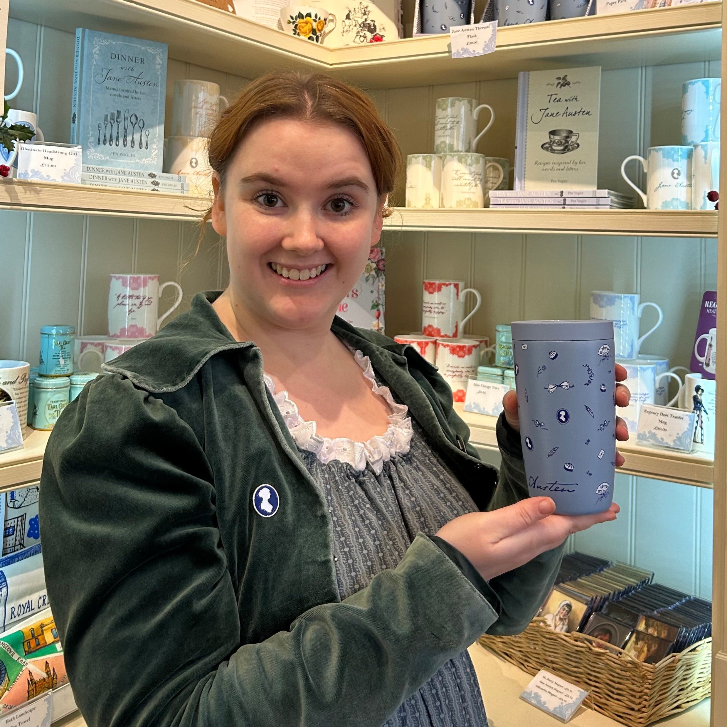 Blue Travel cup model, with icons of a quill, a fan and the famous silhouette of Jane Austen. Modeled by a lady in a gift shop