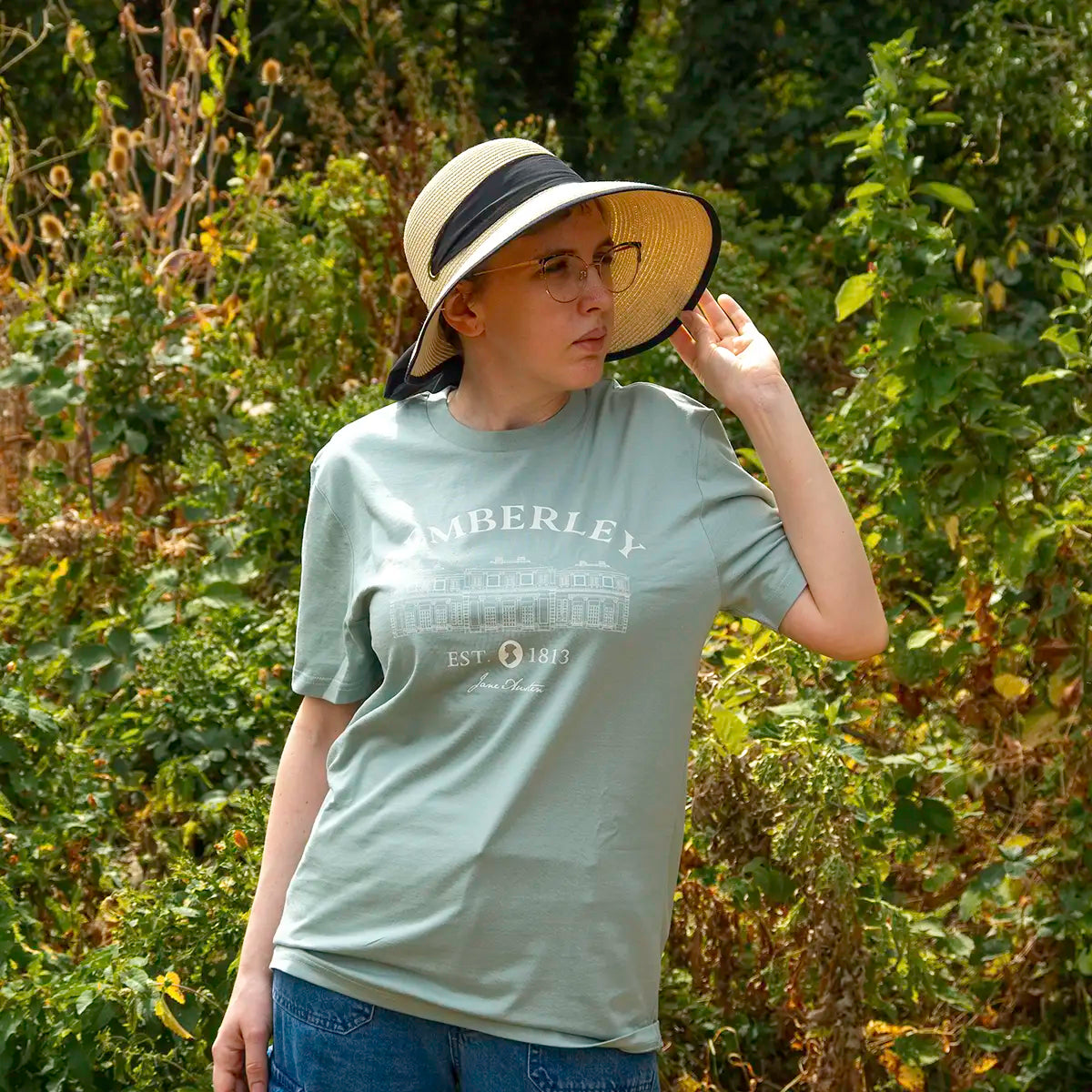 a model standing in front of trees wearing a hat and a Jane Austen Pemberley T-Shirt in green