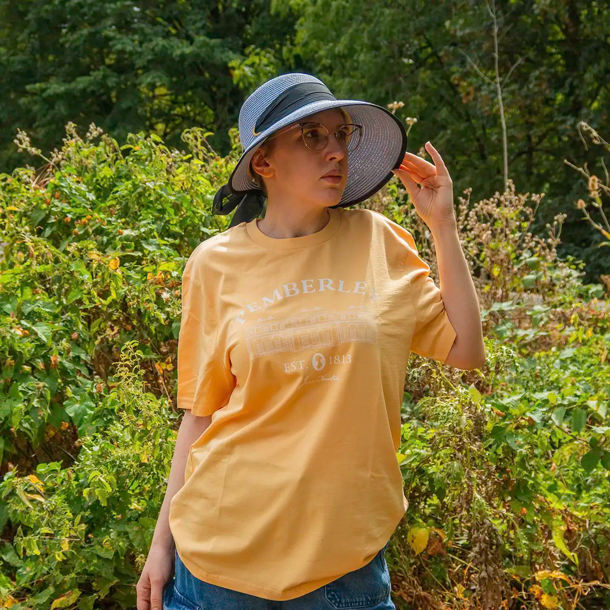 a model in front of a bush and some trees wearing a blue bonnet and a yellow Jane Austen Pemberley T-shirt in size M