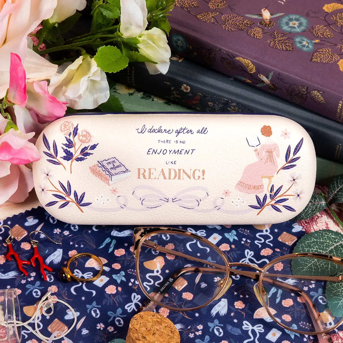 A decorative eyeglasses case with a cream background and whimsical illustrations, including a girl in a pink dress and the phrase 'I declare after all there is no enjoyment like reading!'. The case is styled in a flat-lay arrangement with glasses, flowers, and books.