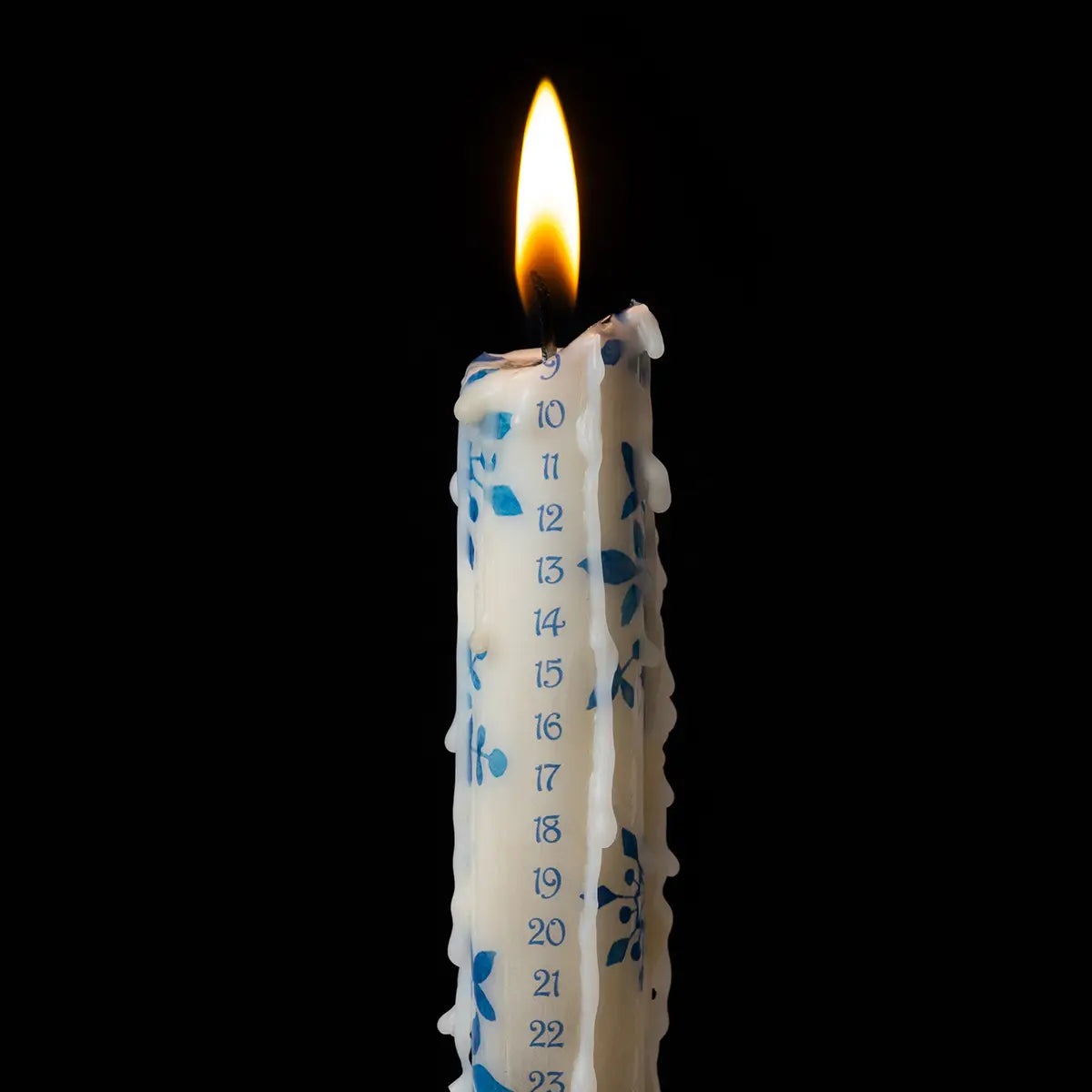 A white advent candle with blue numbers and detailing, lit with wax dribbling down the sides, against a black background