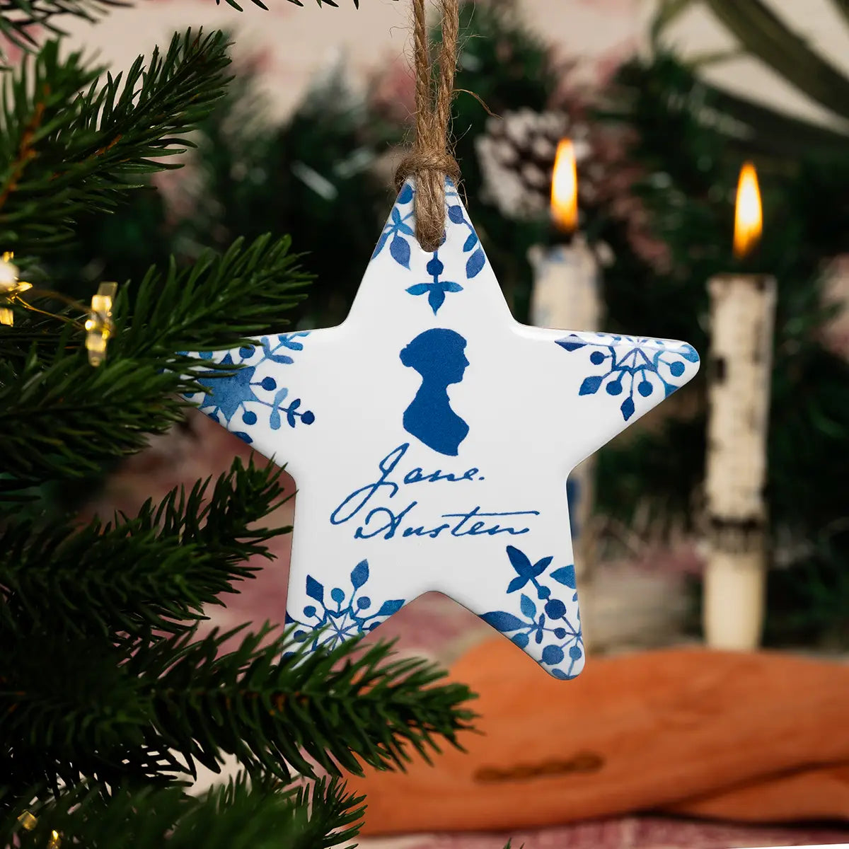 A white ceramic star shaped christmas decoration with a blue Jane Austen themed print, hanging from an artificial christmas tree in front of a christmas tableaux including lit candles, regency gloves, and christmas plants