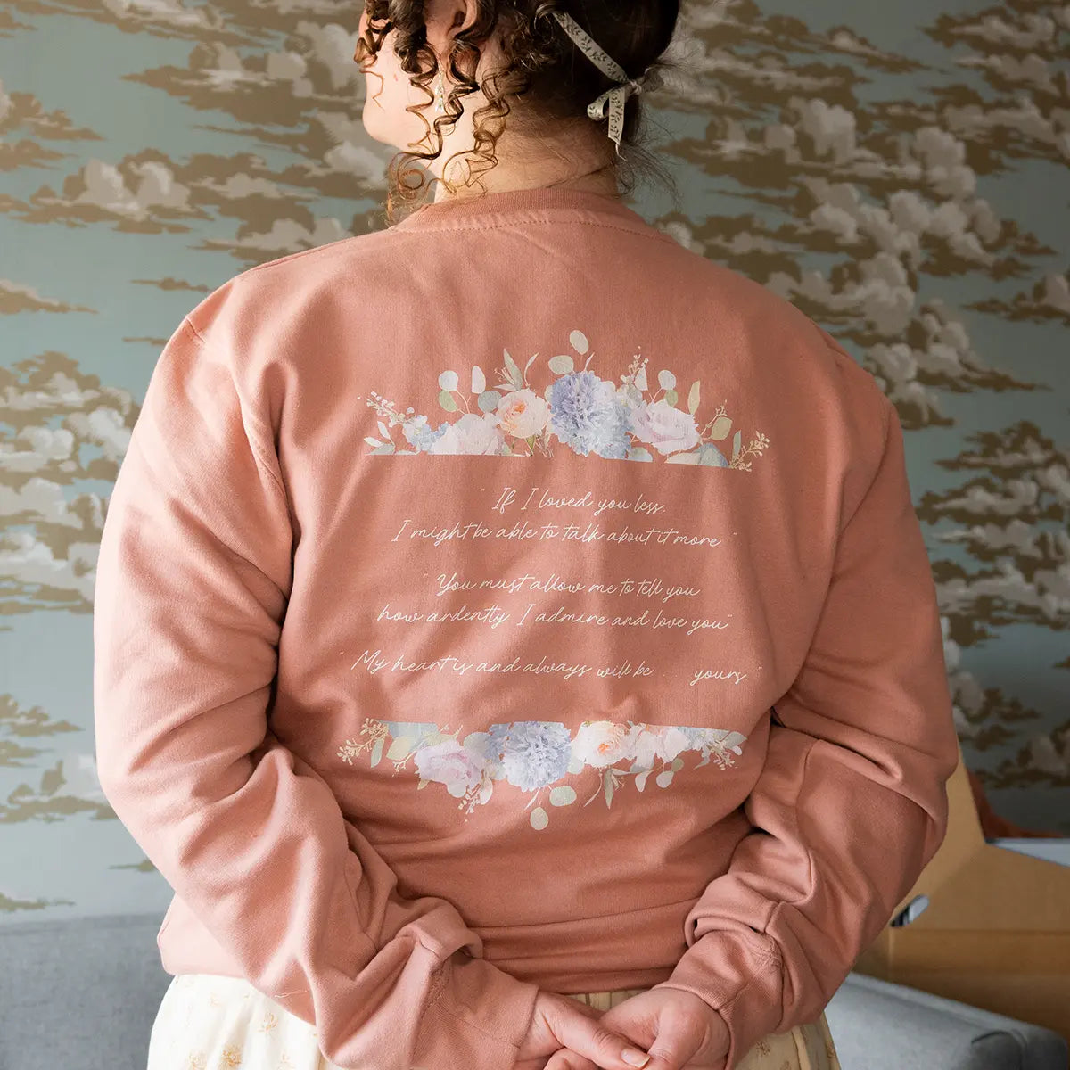 Woman wearing pink Jane Austen themed sweater against blue wall with gold and silver clouds