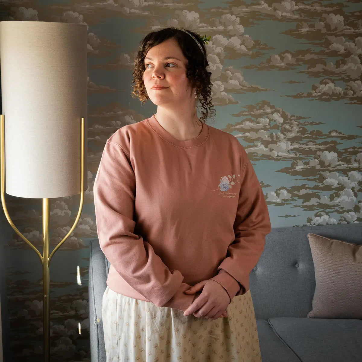 Woman wearing Jane Austen themed sweater in room with lamp, sofa, and cloudy wallpaper
